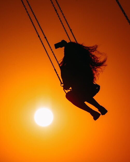 Silhouette woman swinging against orange sky