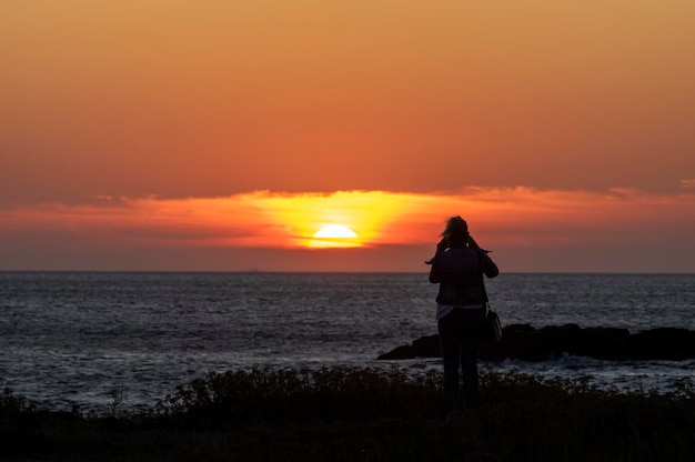 写真 海に沈む夕日のスマートフォンで写真を撮るシルエットの女性