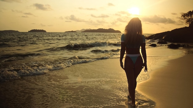 해변 해안선에서 혼자 걷는 실루엣 여성은 따뜻한 금빛 일몰을 휴식을 취합니다. 파란색 수영복과 선글라스를 끼고 물 위를 걷는 여성 개념 휴식 열대 리조트 여행 관광 행복한 여름 휴가