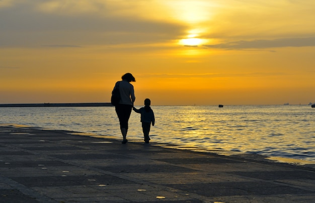 Silhouette di una donna con un bambino, si tengono per mano su uno sfondo di tramonto sul mare