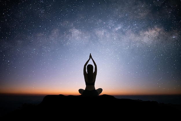 Silhouette of young asian woman practices yoga and meditates on\
top of the mountain with night sky star milky way over the sky she\
likes to practice yoga in the morning it makes her feel calm