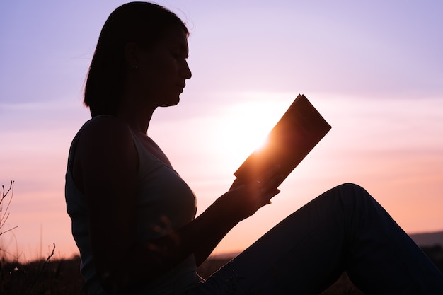 Silhouette of a young beautiful woman at dawn sitting on the ground and carefully staring at the open book