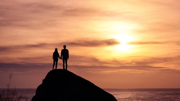 シルエットの若いカップルが熱帯の海の大きな岩に沈む夕日を見ている