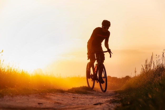 Silhouette young of cyclist on a gravel bike riding on a dust\
trail at sunset