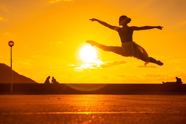 Silhouette of a young female dancer performing a jump along the beach at sunset wearing a tutu