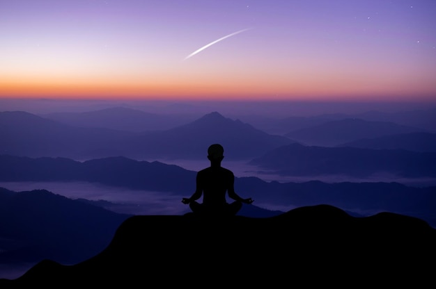 Silhouette of young female sitting practices yoga and\
meditating in lotus position alone on top of the mountain with\
night sky star milky way and meteor she felt calm and happy