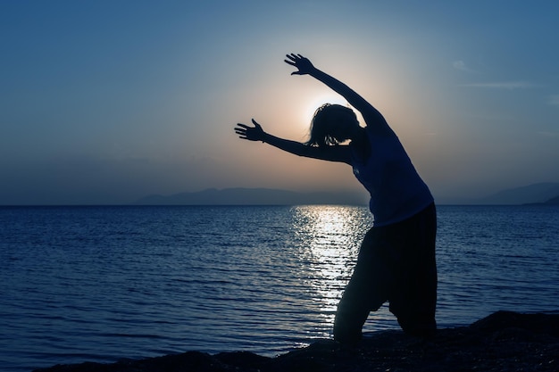 Силуэт молодой девушки, стоящей на пляже с поднятыми руками на закате против солнца