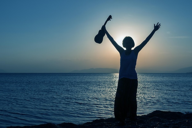 Silhouette of young girl standing on the beach with hands up with ukulele at the sunset against the sun