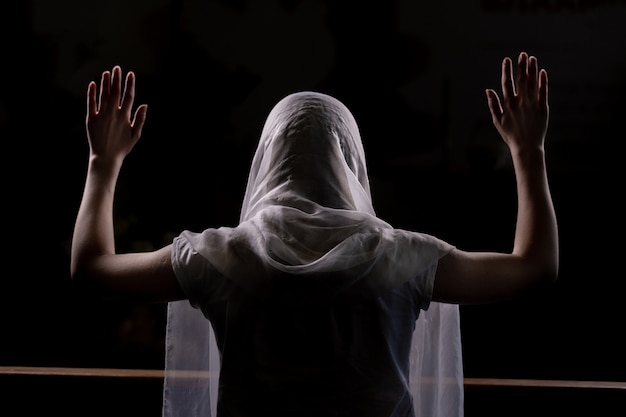 Silhouette of a young girl who sits in church and praying with hands raised up. Close-up view from behind. Backlight