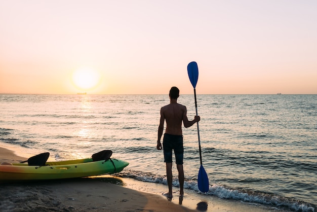 Silhouette of a young handsome guy standing on the beach of the ocean holding a paddle in his hand.