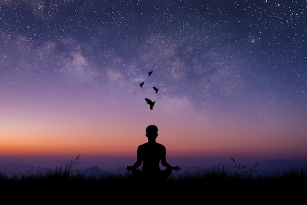 Silhouette of young male sitting practices yoga and meditating\
in lotus position alone on top of the mountain with night sky star\
and milky way he felt calm and happy