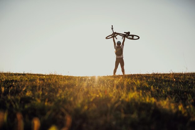 背景に劇的な空と夕日のフィールドで自転車を保持している若い男のシルエット高品質の写真