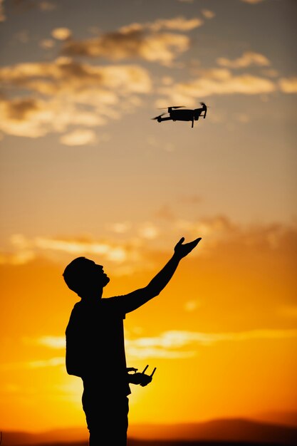 Силуэт молодого человека, управляющего дроном в сельской местности на закате