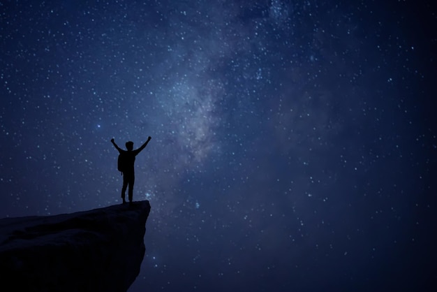 Photo silhouette of young man standing alone on top of mountain and raise both arms enjoying nature on beautiful night sky star milky way background demonstrates hope and freedom