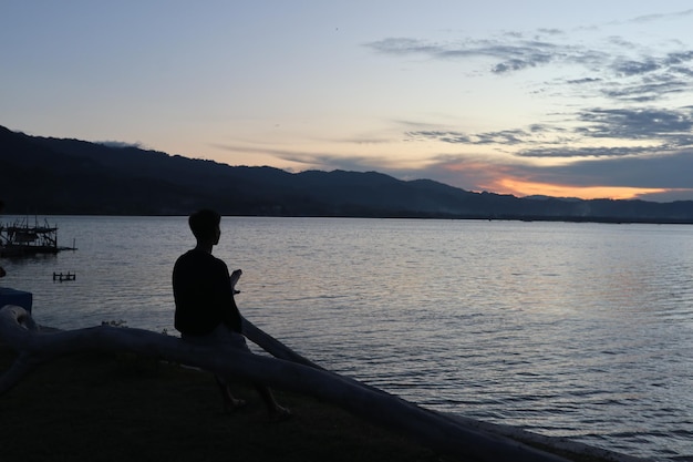 夕日を楽しんでいる湖のそばに立っている若い男のシルエット。自然の中でのどかな雰囲気