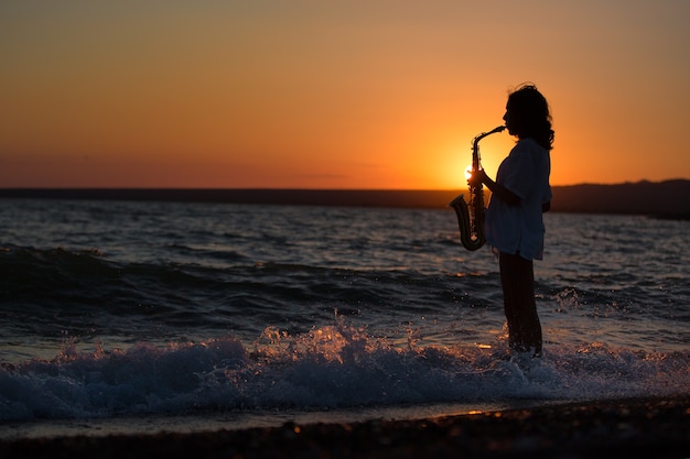 Silhouette of young sexy woman playing saxophone on the beach at sunset