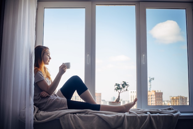 Silhouette of young slender girl sitting on the windowsill at\
home, side view, copy space. outside the window blue sky and tall\
city buildings. red-haired woman holding cup, looking at the\
metropolis.