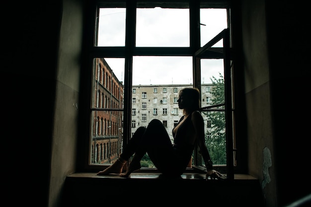 古い家の窓辺に座って誰かを待っている若いスリムな女性のシルエット。