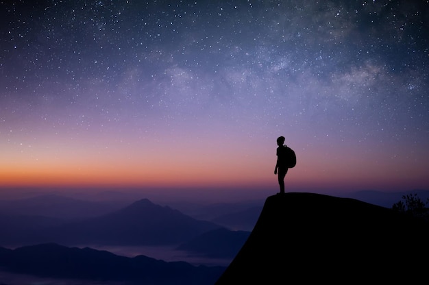 Silhouette of young traveler with backpack standing alone on\
top of the mountain and watched the beautiful view night sky star\
and milky way he was happy to travel alone in the wide world