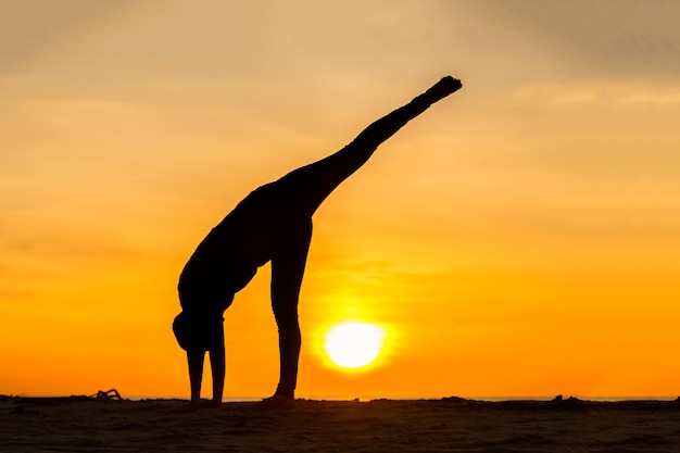 Silhouette young woman practicing yoga on the beach at sunset.