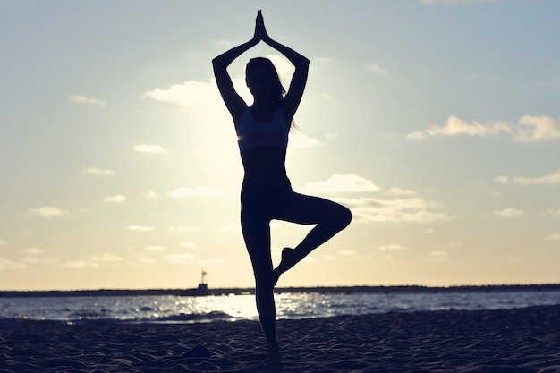 silhouette young woman practicing yoga on the beach at sunset