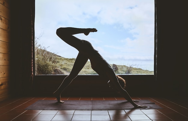 Silhouette di giovane donna che pratica yoga indoor sullo sfondo di una grande finestra con paesaggio naturale