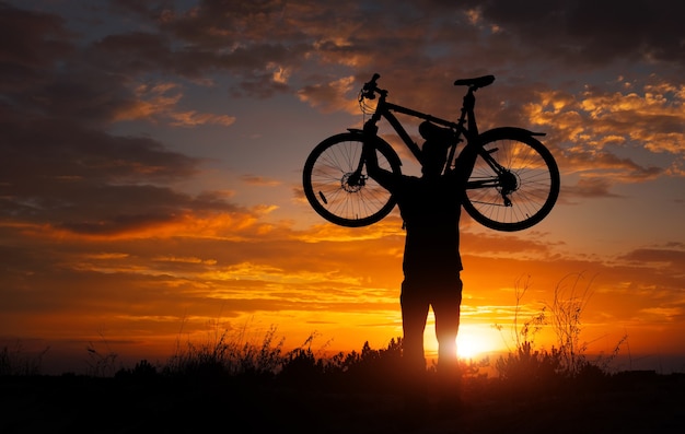 Silhouetteer de man die in actie staat met het optillen van de fiets boven zijn hoofd op de weide met zonsondergang