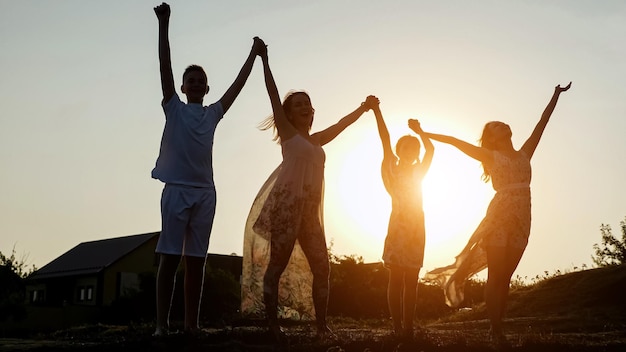 Silhouetten van een vrolijke moeder met kinderen die handen opsteken die op de dorpsweg staan onder de heldere hemel bij het achterlicht van de zonsondergang in de zomeravond