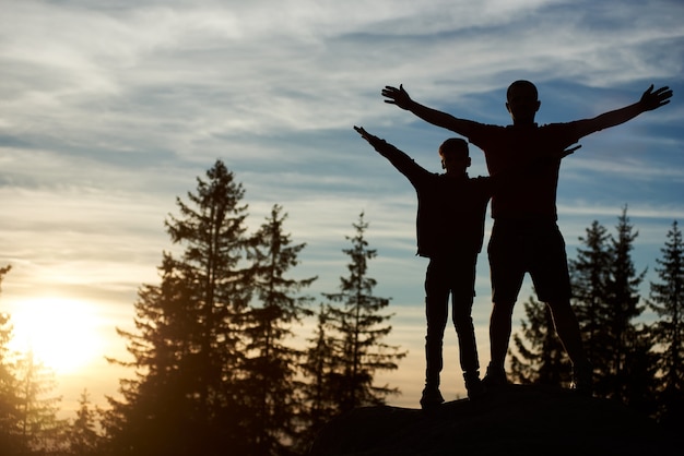 Silhouetten van gelukkige vader en zoon die 's avonds op de top van de berg staan