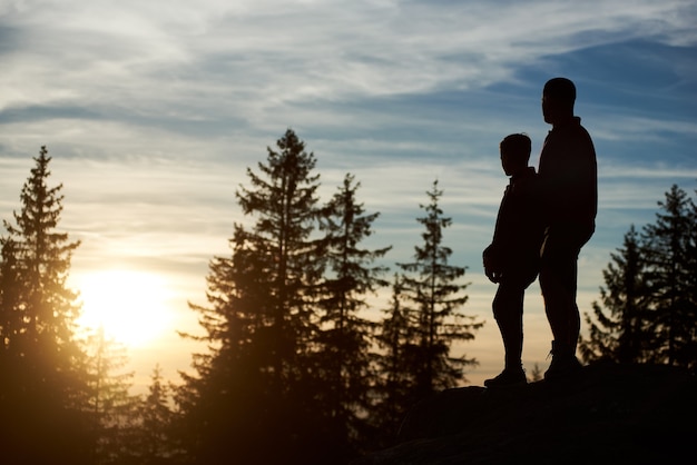 Silhouetten van jonge vader en zoon die 's avonds op de top van de berg staan
