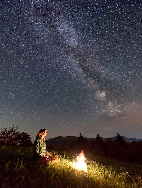 Silhouettes of girl near haystack and bonfire under starry sky on which Milky Way is visible