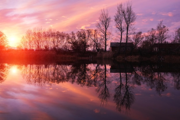 明るい緋色の秋の夕日を背景に、湖の放棄された村の家や木のシルエット。