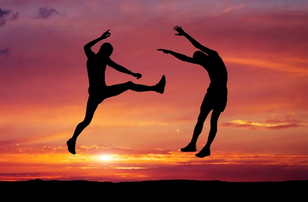 Silhouettes of two fighters on sunset fiery background. Battle at sunset. Kick in the air at the opponent's body.