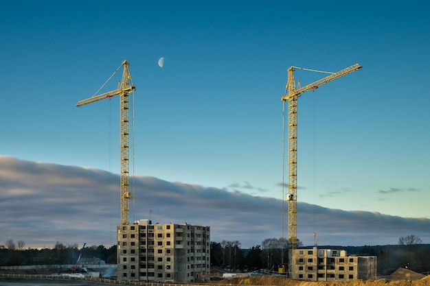 Silhouettorenkranen en onvoltooide hoge gebouwen met meerdere verdiepingen in aanbouw in bewolkte dag met maan