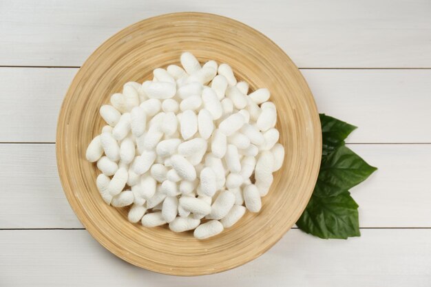 Silk cocoons with plate and mulberry leaves on white wooden table flat lay