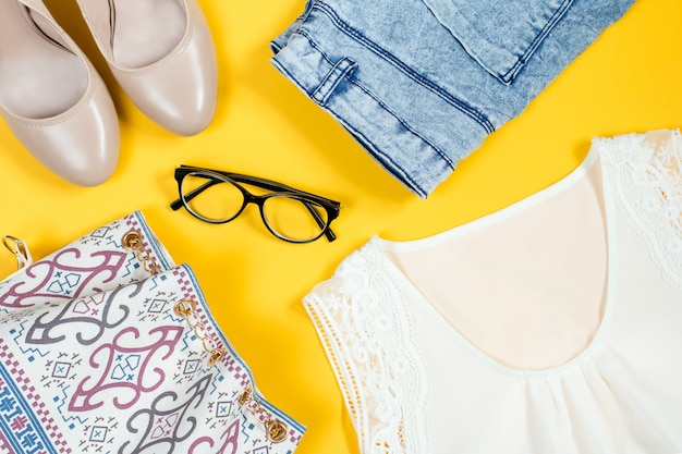Silk white top, denim shorts, nude shoes, purse, black eyeglasses on a bright background