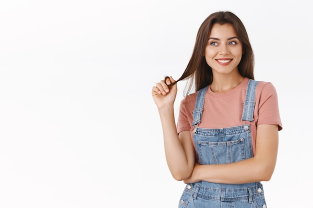 Sciocco, sognante seducente donna bruna in salopette, t-shirt che rotola la ciocca di capelli sul dito e sorride felice, distogliendo lo sguardo come ricordando, immaginando qualcosa di buono, in piedi sfondo bianco soddisfatto