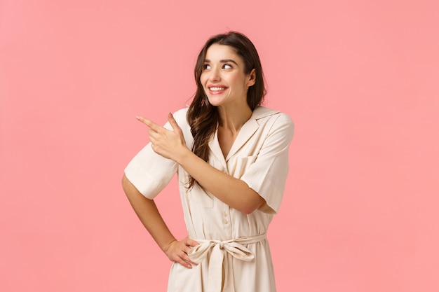 Silly girl anticipating delivery cute new dress for prom. Cheerful european brunette woman in dress eager try out product, pointing upper left corner, smiling and laughing, standing pink wall