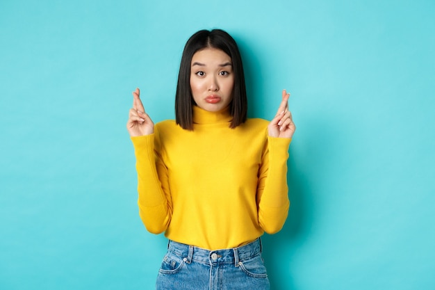 Silly hopeful asian girl making wish, pucker lips and looking at camera with dreamy glance, cross fingers for good luck, standing over blue background.