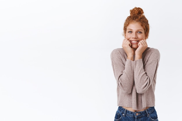 Silly, lovely caucasian redhead woman with curly hair, showing white beautiful smile, squeez cheeks and gazing camera with affection, admiration and happiness, white wall