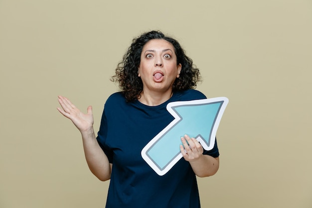 Silly middleaged woman wearing tshirt holding arrow mark pointing to side looking at camera showing tongue and empty hand isolated on olive green background