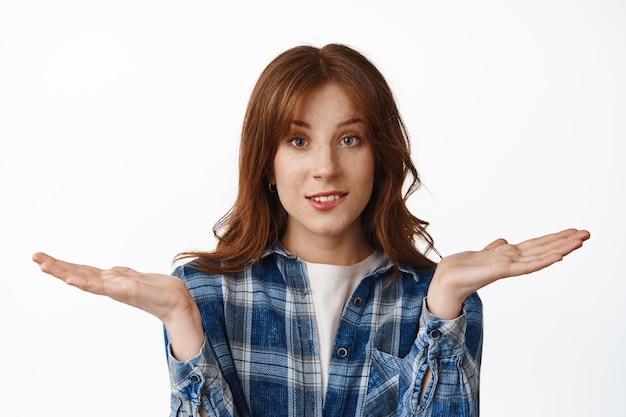 Silly redhead girl shrugging shoulders and bite lip, dont know nothing, have no idea, standing clueless and indecisive, puzzled what to do, standing against white background