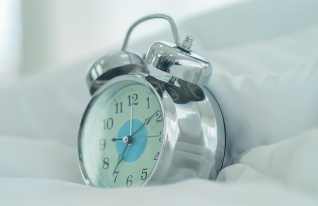 Silver chrome vintage alarm clock placing on a white bed pillow\
sheet for waking up in the morning concept