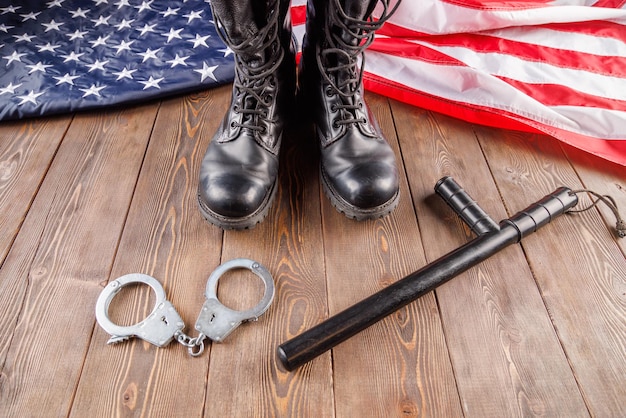 Silver metal handcuffs black ankle boots and police nightstick near US flag on wooden surface