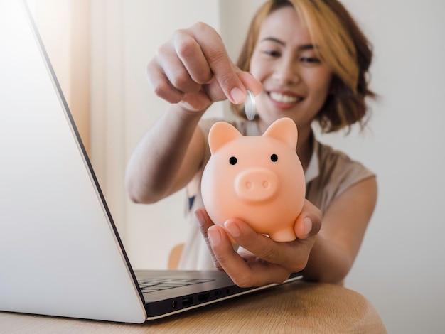 Foto moneta d'argento che mette nel salvadanaio rosa nella mano della donna asiatica con un sorriso felice mentre si utilizza un computer portatile sulla scrivania in legno i risparmi di tecnologia di investimento donano concetti online