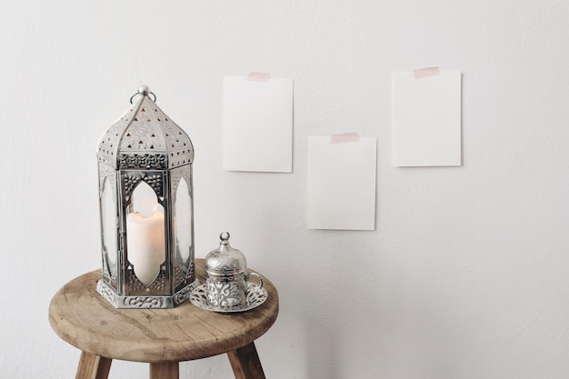 Silver Moroccan Arabic lantern with cup of Turkish tea coffee drink on old wooden stool Blank greeting card mockups taped on white wall Muslim holiday Ramadan Kareem Festive background