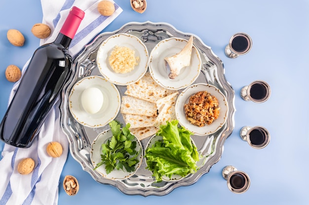 Серебряное блюдо с традиционными угощениями к еврейской пасхе, бутылка красного кошерного вина и налитые серебряные бокалы, вид сверху на синем фоне