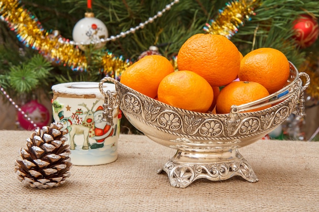 Silver vase with oranges, a candle and a cone on sackcloth. Christmas fir tree with toy balls and garlands on the background. Christmas Eve concept.