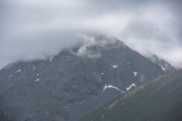 山腹に霧のかかった森と灰色の低い雲の間に高い山がある、シンプルな大気の雨の風景灰色の曇り空の下に山がある荒涼とした曇りの風景暗い高山の航空写真