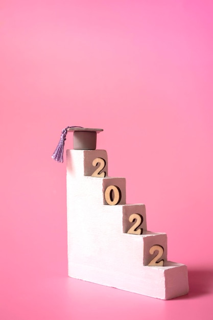 Semplice concetto di educazione su sfondo rosa graduazione 2022 gradini del podio con tappo di graduazione e numeri in legno primo piano formato verticale